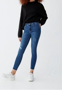 Persona con un suéter de punto negro, jeans ajustados desgastados azules y zapatillas blancas, de pie con una mano en el bolsillo contra un fondo blanco.