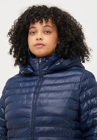 Marineblaue Steppjacke mit hohem Kragen, Reißverschluss und Kapuze. Besitzt ein horizontales Steppmuster und ein kleines Logo auf der Brust.
