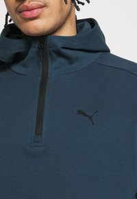 Felpa con cappuccio blu scuro dotata di un alto colletto, chiusura a zip e logo Puma nero. Realizzata in un morbido tessuto texturizzato con un design sportivo.