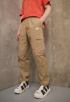 Personne portant un pantalon cargo Nike beige, une chemise rouge et des baskets blanches Nike Air, debout devant un mur beige texturé.