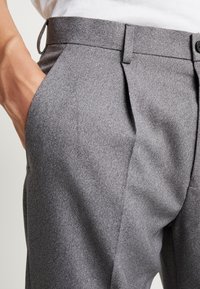 Grå skräddarsydda byxor med texturerad yta, sidofickor och platt framdesign. Bälteshällor syns vid midjan.