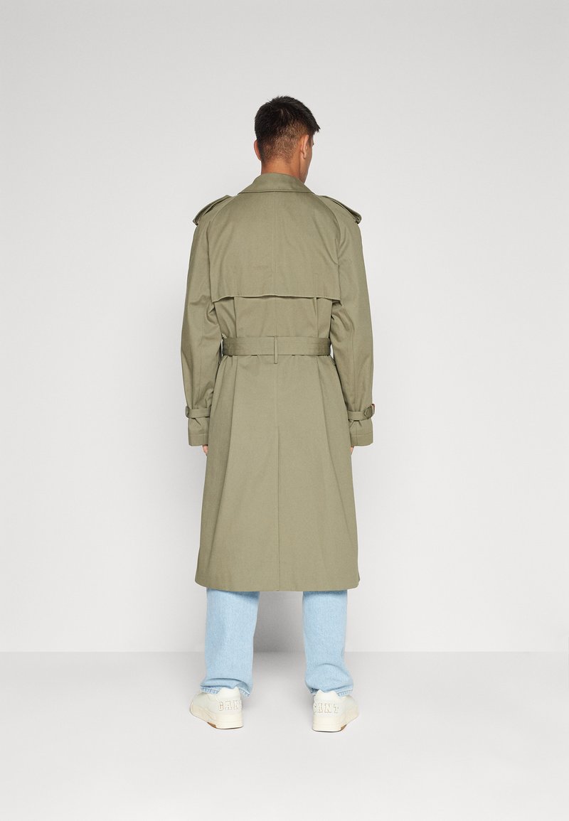 Olivgrüner Trenchcoat mit Schulterklappen, Gürtel in der Taille und Schulterklappen, kombiniert mit hellblauen Jeans und cremefarbenen Sneakers.