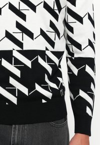 Schwarzer und weißer geometrischer Musterpullover mit langen Ärmeln, getragen mit dunkelgrauen Jeans, zeigt den mittleren Oberkörper und den rechten Arm.