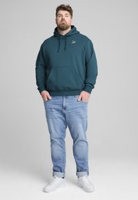 Felpa teal con tasca a marsupio, cappuccio regolabile con cordino e logo. Indossata con jeans azzurri e sneakers bianche. Tessuto morbido e casual.