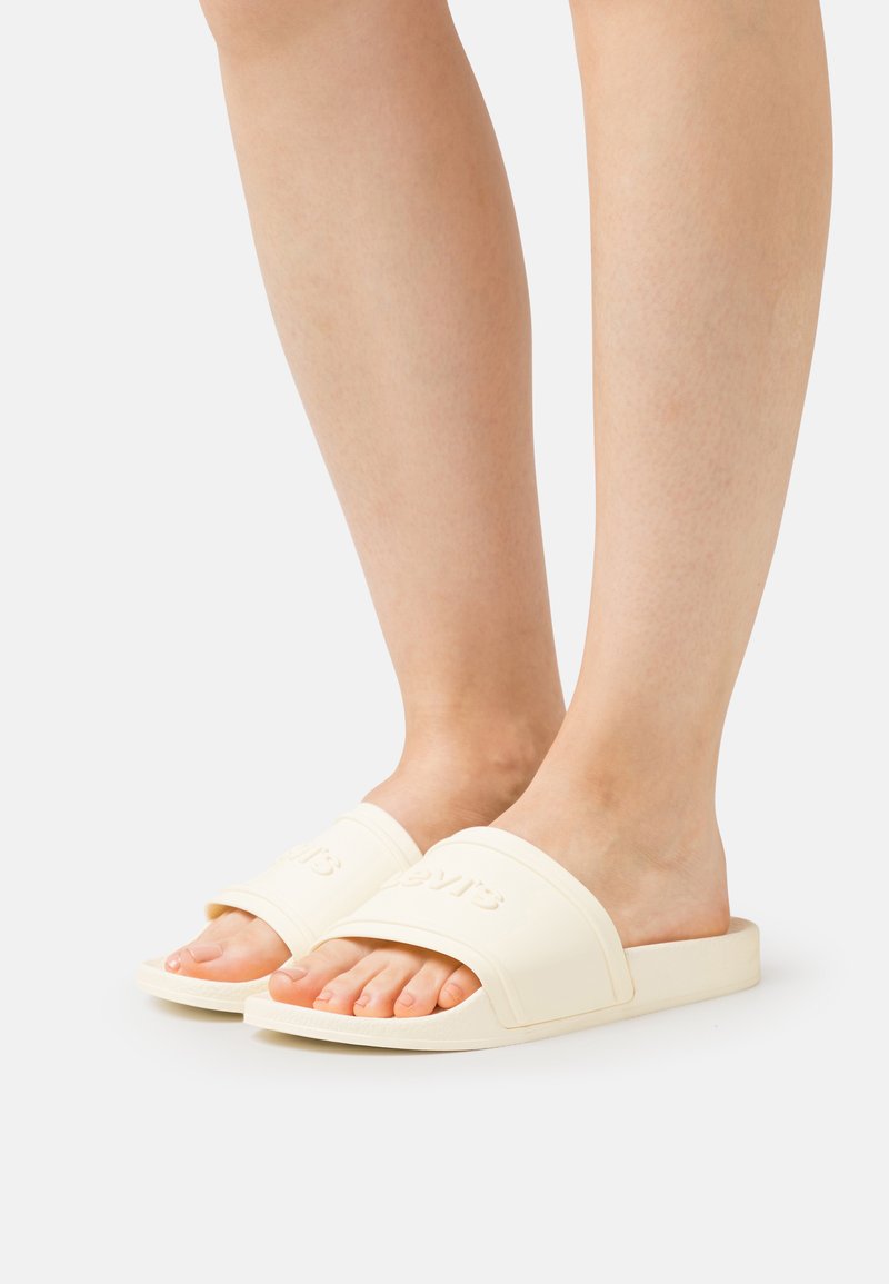 Helle beigefarbene Riemchensandalen aus weichem Gummi, mit einem breiten Riemen mit geprägtem Logo, offenem Zehendesign und flachen Sohlen für zusätzlichen Komfort.