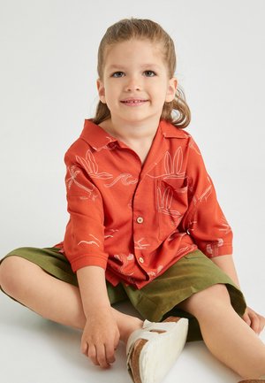 Camisa corta de manga naranja con patrones botánicos blancos, cierre de botones y bolsillo, combinada con pantalones cortos verdes oliva. Niño sentado en una superficie plana.