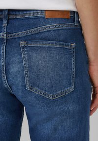 Dżinsy z denimu w ciemnym niebieskim kolorze, z tylną kieszenią z zaostrzoną klapką, pomarańczowym przeszyciem i brązową skórzaną metką marki nad paskiem talii.