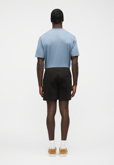 Homme portant une chemise bleu clair à manches courtes, un short noir, des chaussettes blanches et des baskets beige, debout de dos sur un fond uni.