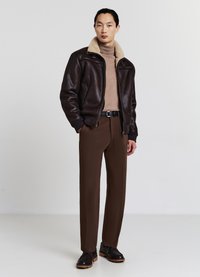 Giubbotto bomber in pelle marrone con colletto in shearling beige, abbinato a pantaloni marroni e scarpe nere, caratterizzato da un design elegante e aderente.