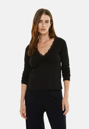 Mujer con cabello largo y castaño vistiendo un top negro de manga larga con escote en V adornado con encaje y pantalones negros, posando frente a un fondo liso.