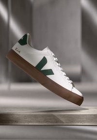 Valkoiset tennarit, joissa on vihreät logo-yksityiskohdat ja ruskea kumipohja. Ominaisuuksiin kuuluu sileä pinta, pyöreä muoto ja tasaiset nauhat. Minimalistinen muotoilu.