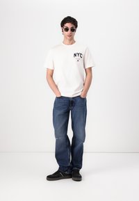 White short-sleeve t-shirt with "NYC" printed on the chest, paired with blue denim jeans. Black sneakers and dark sunglasses complete the look.