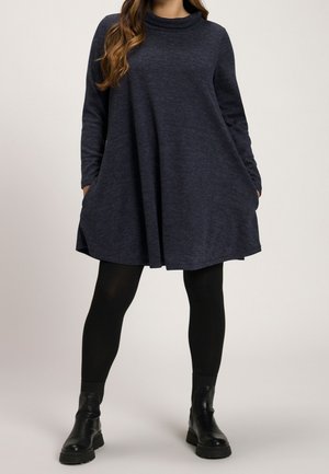 Femme portant une robe ample à manches longues gris foncé avec des poches, des leggings noirs et des bottines noires, debout devant un fond uni.