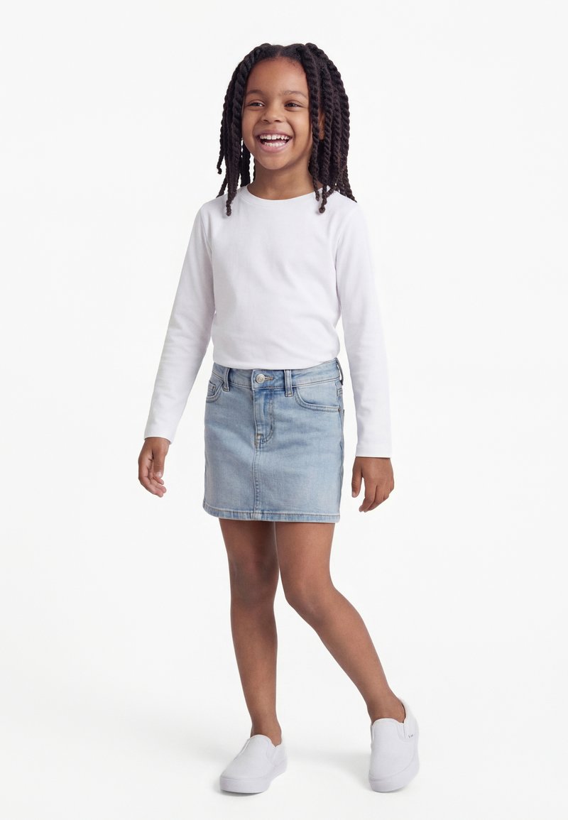 Šypsanti jauna mergina su pintais plaukais, dėvinti baltą ilgomis rankovėmis marškinėlį, šviesios džinsinės spalvos sijoną ir baltus slip-on tipo batelius, stovinti ant balto fono.