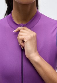 Osoba o średnim odcieniu skóry zaciągająca zamek błyskawiczny na fioletowym, bezrękawnym ubraniu z wysokim kołnierzem.