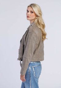 Lichtgrijze suède leren jas met ritssluiting aan de voorkant, cropped ontwerp en schouderepauletten, gecombineerd met denim jeans met metalen accenten.