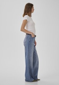 Ženska, postavljena v profilu, nosi oprijeto belo majico, modre kavbojke širokih hlačnic z visokim pasom in čevlje s koničastim prstom, na enobarvnem ozadju.