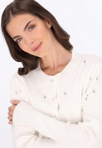 Biały kardigan z prążkowanej dzianiny, ozdobiony perłowymi guzikami i srebrnymi koralikami na ramionach. Długie rękawy z wydłużonymi mankietami, miękka tekstura.