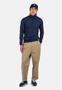 Marineblauer Pullover mit Reißverschluss am Kragen, beigen, lockeren Hosen und marineblauen Sneakers. Der Pullover hat eine strukturierte Oberfläche und einen kontrastierenden roten Kragen.
