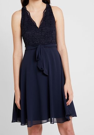 Ærmeløs navyblå kjole med blondeudskæring i V-hals, stofbælte i taljen og flydende chiffonnederdel, der ender over knæene.