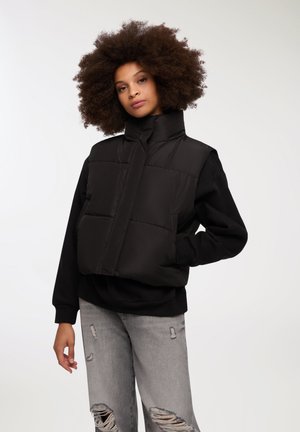 Gilet piumino nero con colletto alto, pannelli rettangolari e tasche frontali, indossato sopra una felpa nera. Modello abbinato con jeans grigi scuciti.