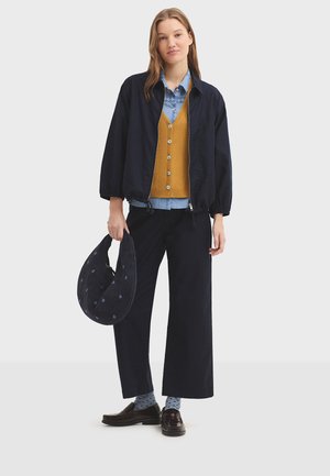 Young woman wearing navy jacket, mustard cardigan, blue shirt, wide-leg pants, polka-dot socks, loafers, holding a navy handbag.