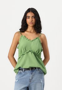 Grønn, ermester topp med rynkede detaljer, teksturert stoff og V-hals. Kombinert med lyseblå, revne jeans og et svart belte.