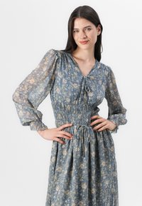 En kvinna med långt mörkt hår har på sig en blå blommig klänning, ler och poserar med händerna på höfterna mot en enkel vit bakgrund.