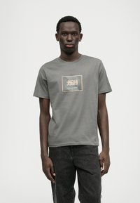 Šedé bavlnené tričko s okrúhlym výstrihom a krátkymi rukávmi, s grafickým logom v bielej a oranžovej farbe na štvorcovom pozadí.