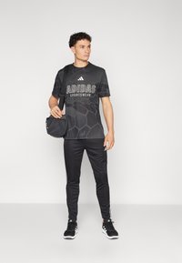 Camisola desportiva preta da adidas com textura geométrica e logótipo, combinada com calças de treino pretas com riscas brancas laterais e sapatilhas pretas.