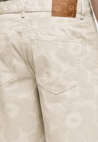 Μπεζ παντελόνι με διακριτικό μεγάλο λουλουδάτο σχέδιο, ορατές πίσω τσέπες, θηλιές για ζώνη και καφέ δερμάτινο ταμπελάκι με ανάγλυφο σχέδιο λουλουδιού.