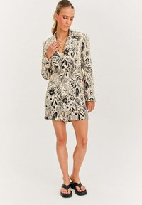 Kratká rukávová blézer šaty v krémové barvě s černými organickými vzory. Obsahuje páskový pas, klopy a volný střih. Nosené s černými žabkami.