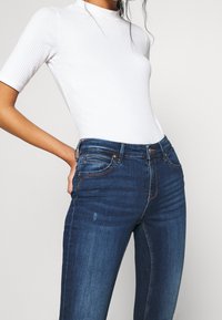 Vitt ribbat bodysuit med korta ärmar, parat med mörkblå denimjeans som har femfickorsdesign och synliga sömnadsdetaljer.