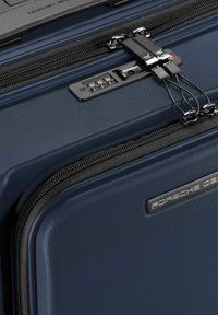 Maleta de carcasa dura azul marino con superficie texturizada, doble cremallera, cerradura de combinación y un logotipo plateado de "Porsche Design" en el lateral.