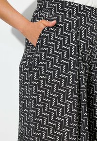 Ruka s opálenou pletí odpočívá v kapse kalhot s černo-bílým zigzag vzorem na bílém pozadí.