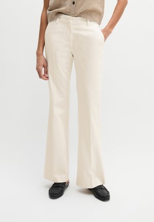Personne portant un pantalon large crème avec poches avant, associé à des mocassins noirs sans lacets et un haut en maille beige.