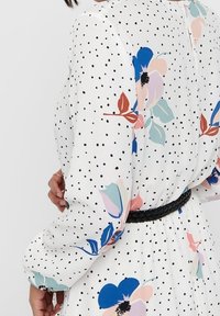 Nainen valkoisessa mekossa, jossa on mustia pilkkuja ja värikkäitä kukkakuoseja, käyttää vyötäröllään mustaa punottua vyötä.