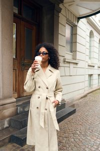 Trench coat bej cu două rânduri de nasturi, cu curea, nasturi mari și mâneci lungi. Persoana poartă o ceașcă de cafea și stă pe trepte de piatră cubică.