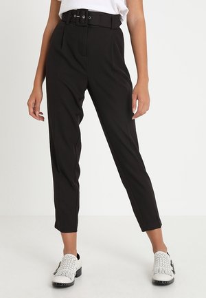 Femme portant un pantalon noir taille haute fuselé avec une large ceinture et des mocassins blancs perforés sur un fond blanc uni.