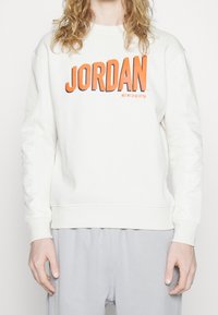 Sweat-shirt blanc avec "JORDAN" imprimé en lettres orange vif. Caractérisé par une texture douce et des poignets côtelés, accompagné d'un pantalon de jogging gris clair.