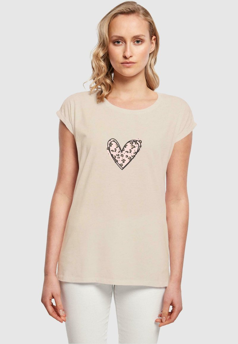T-shirt beige a maniche corte realizzato in tessuto morbido, con un design a forma di cuore con stampa leopardata rosa al centro. Collo rotondo classico e vestibilità rilassata.