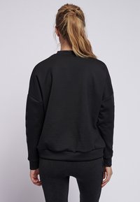 Svart sweatshirt med översized passform, ribbade mudd och nederkant. Slät textur med subtila logotyper på båda ärmarna. Bärs med matchande mörka leggings.