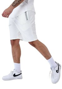 Pantalones cortos deportivos blancos con un diseño elegante, que cuentan con un bolsillo lateral y acentos grises. Combinados con calcetines blancos y zapatillas Nike negras.