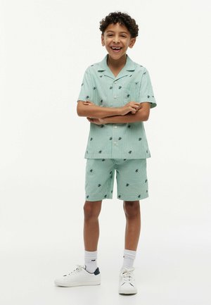 Junge mit lockigem Haar, der ein mintgrünes gestreiftes Kurzarmhemd und passende Shorts trägt, mit verschränkten Armen steht und lächelt.