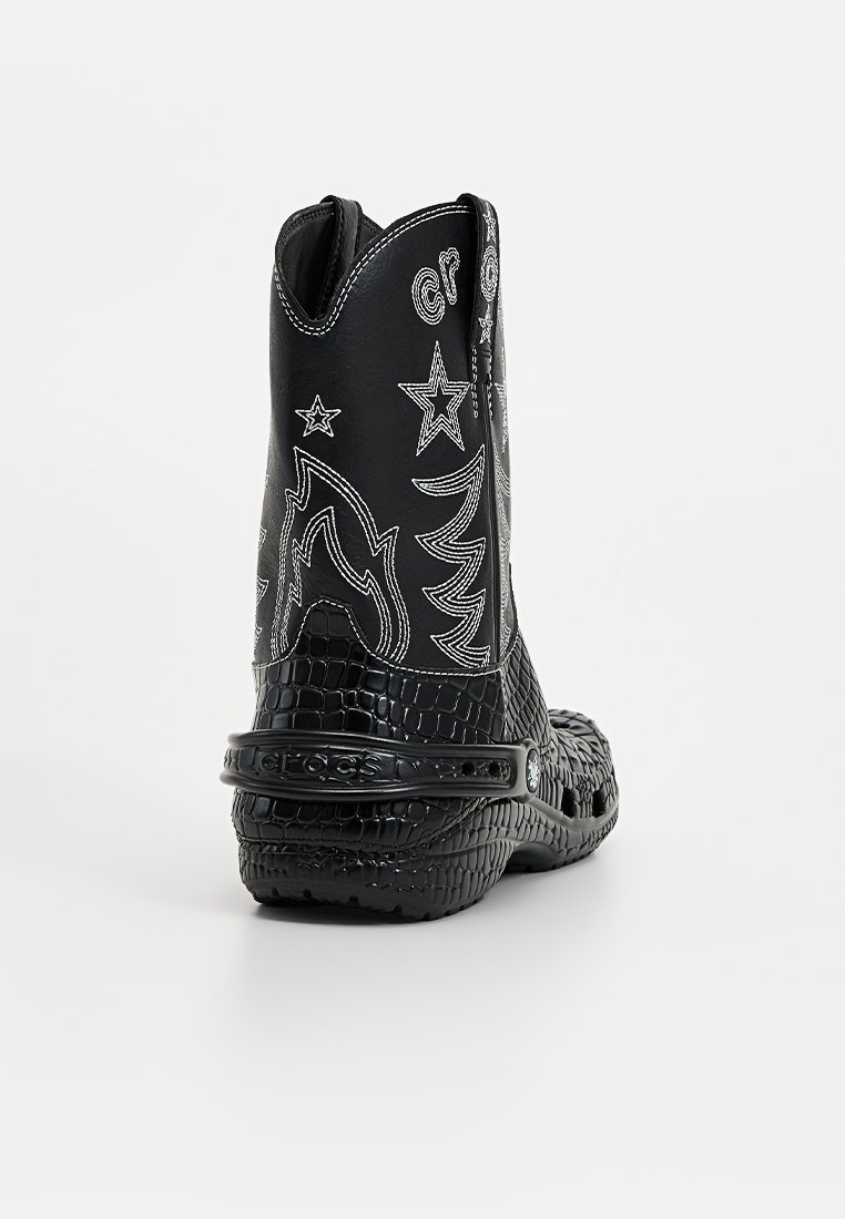 Botas negras de tobillo con textura de cocodrilo, estrellas y diseños bordados en blanco, y una correa en el talón para mayor soporte.