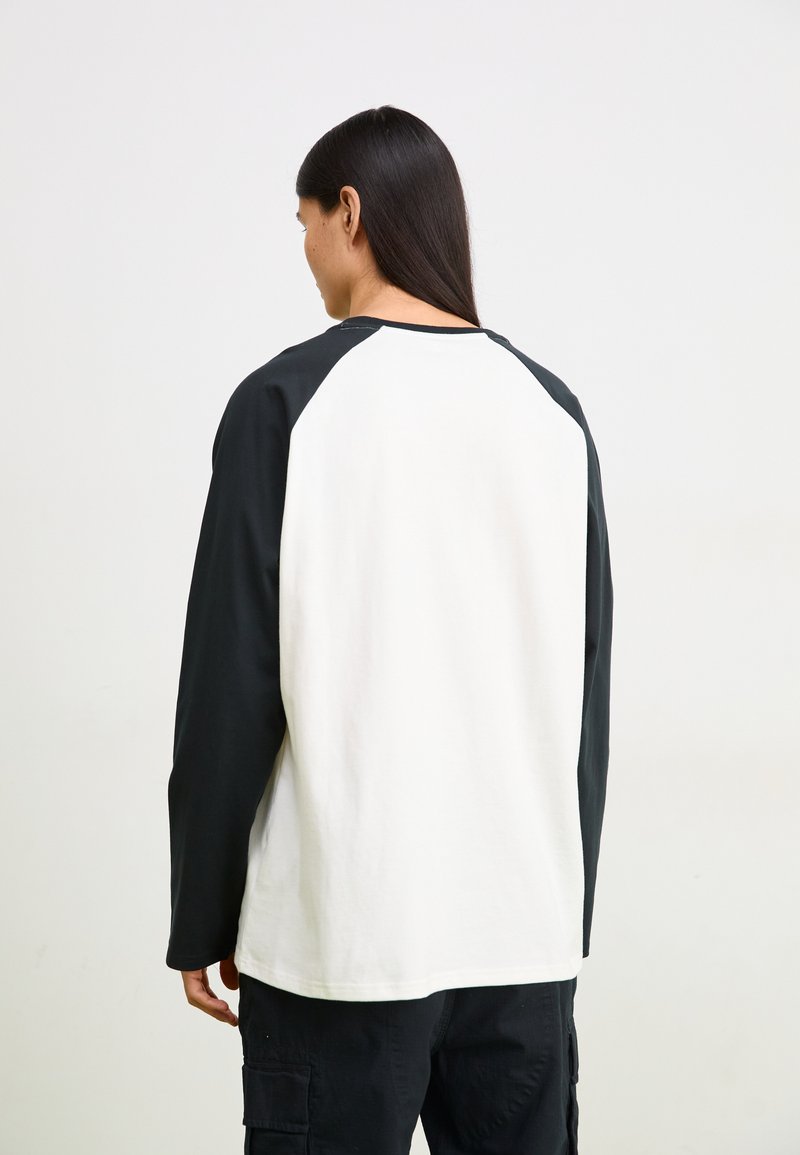 Chemise à manches longues avec des manches raglan noires et un corps blanc. Matériau en coton, col rond et détails de couture visibles.