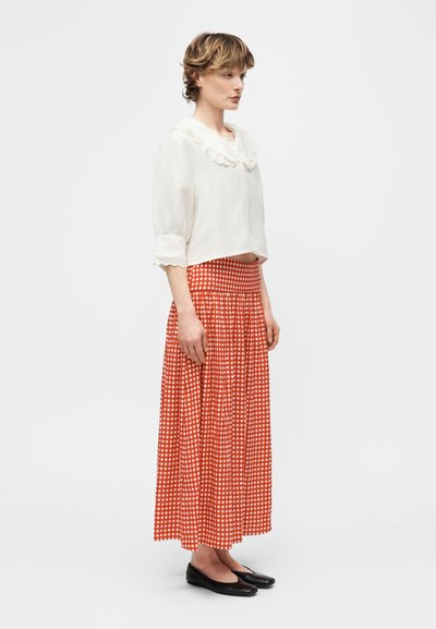 Γυναίκα με κοντά μαλλιά που φοράει λευκό πουκάμισο με βολάν στον γιακά και τα μανίκια, μακριά κόκκινη και λευκή καρό φούστα και μαύρα παπούτσια, στέκεται πλαγίως.