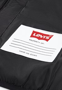 Crni materijal s bijelom etiketom koja sadrži "Levi's" logotip, polje "VLASNIŠTVO:" i tekst "PRENOSIMO", sa crvenim detaljima.