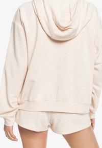 Visuale posteriore di una persona che indossa un felpa con cappuccio color crema e pantaloni corti abbinati, con le mani rilassate ai lati.