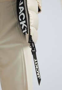 Pudset beige jakke med en sort lynlås og en rem mærket "JACK1T." Stoffet har en glat tekstur og et puffet design.